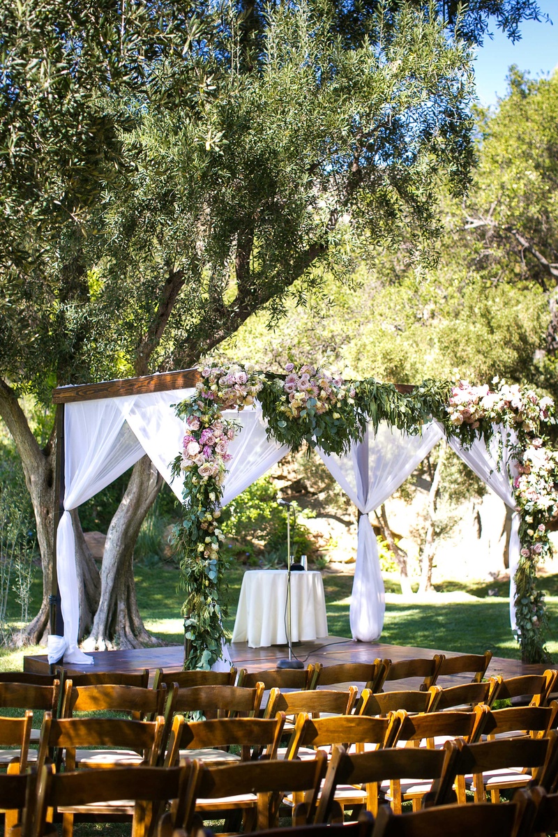 Chuppah with Greenery and Flowers, Sheer Fabric
Photography: Callaway Gable
Read More:
http://www.insideweddings.com/weddings/alfresco-summer-wedding-with-lush-foliage-accents-in-california/805/ A wooden chuppah featuring foliage with light colored flowers and sheer white fabrics hanging