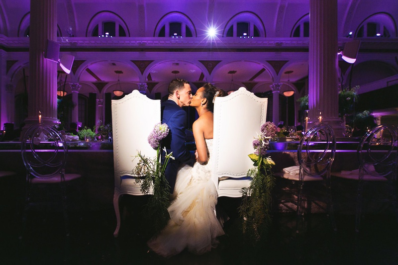 Couple Kiss at Reception Table
Photography: Callaway Gable
Read More:
http://www.insideweddings.com/weddings/modern-in-the-round-ceremony-whimsical-garden-inspired-reception/1155/ Bride and groom kiss at tall chairs with fresh flowers purple lighting kiss reception