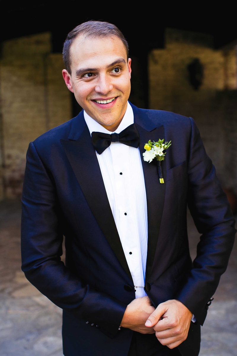 Groom in Dark Blue Tuxedo
Photography: Callaway Gable
Read More:
http://www.insideweddings.com/weddings/alfresco-summer-wedding-with-lush-foliage-accents-in-california/805/ a groom smiling in a dark blue tuxedo and black bow tie with green and white boutonniere