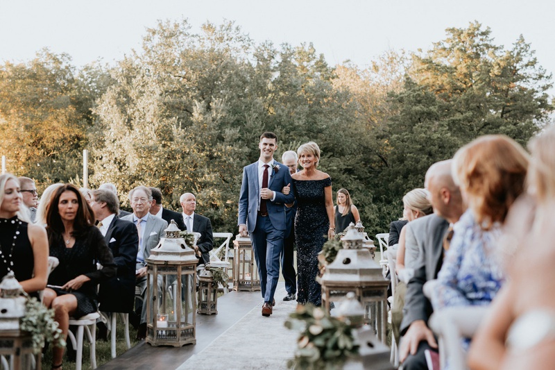 Mother of Groom in Sequin Black Dress
Photography: Grant Daniels Photography
Read More:
http://www.insideweddings.com/weddings/outdoor-ceremony-tented-reception-with-cozy-fall-color-palette/1183/ groom walking mother of groom down aisle off shoulder black sequin dress tall lanterns on aisle