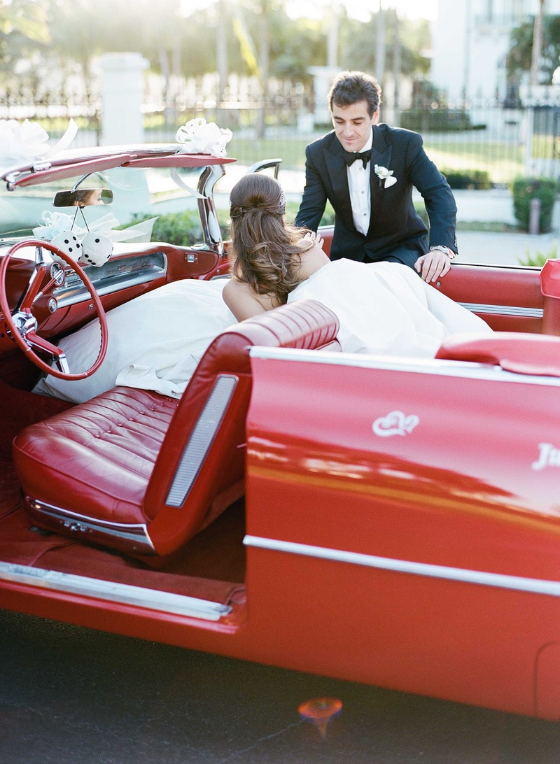 Bride Exits Classic Red Car
Photography: Kat Braman Photography
Read More:
http://www.insideweddings.com/weddings/a-sophisticated-museum-wedding-with-modern-details-in-palm-beach/899/ Groom in tuxedo helping bride out of classic red car with vintage seats and dice die hanging mirror