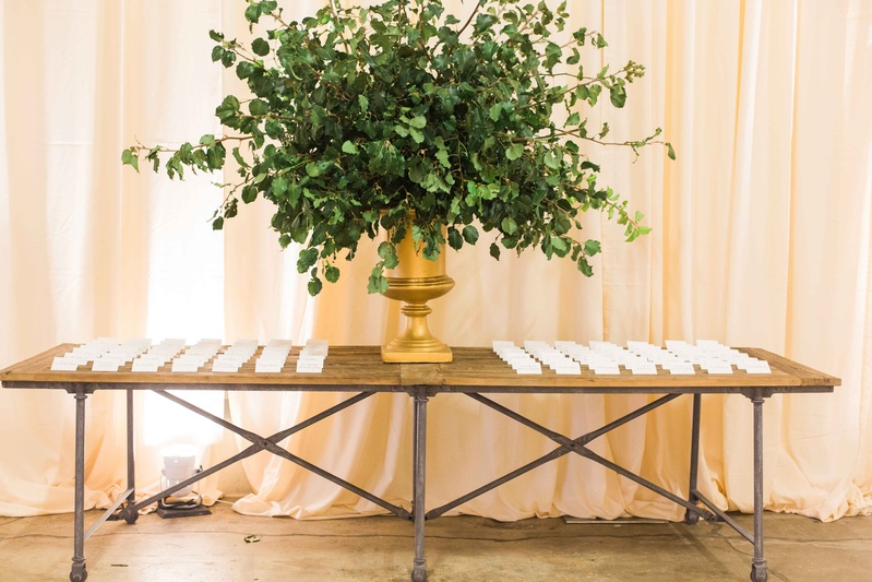 Gold Vessel with Greenery at Escort Card Table
Photography: Gabe Aceves Photography
Read More:
http://www.insideweddings.com/weddings/warm-cozy-wedding-at-unique-warehouse-venue-in-washington-dc/902/ Bare wood table industrial with gold urn filled with green leaves white escort cards on top