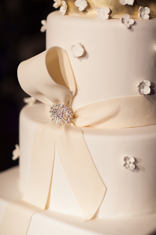 Ribbon %26 Brooch Detail on Cake
Photography: Jay Lawrence Goldman Photography
Read More:
http://www.insideweddings.com/weddings/elegant-white-lavender-ballroom-wedding-with-cherry-blossom-trees/710/ White wedding cake with top golden layer, fondant ribbon, and rhinestone pin, small fondant flowers